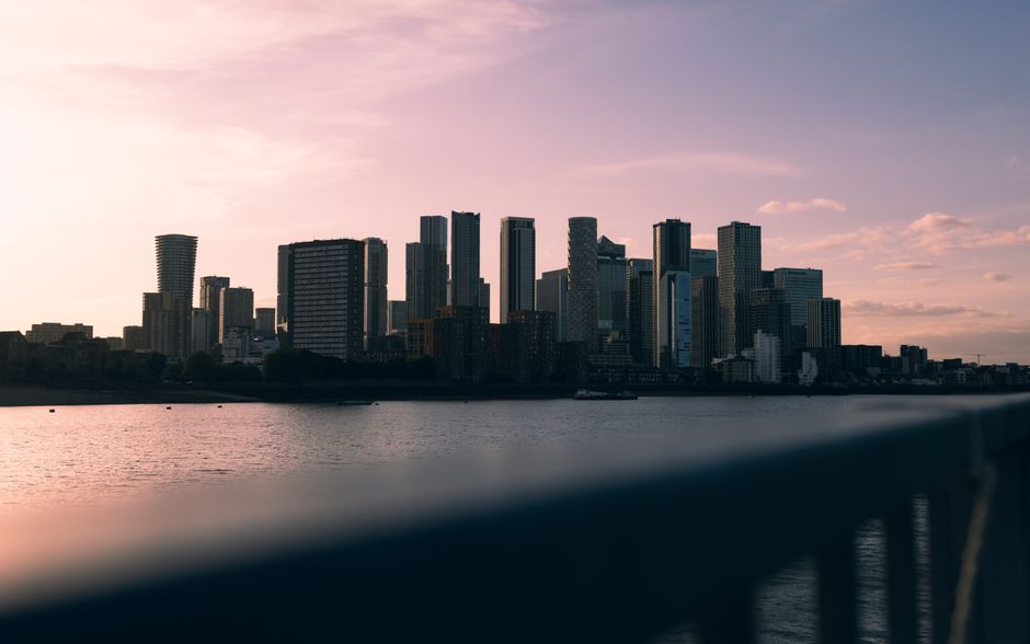 A broad view of Canary Wharf’s skyline at dusk, with modern high-rise office buildings and residential apartments illuminated by interior and exterior lights. The buildings are situated along the wa
