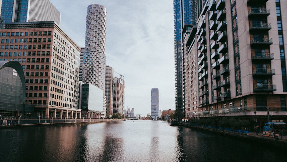 A panoramic view of Canary Wharf in London, featuring a canal running through the city with tall modern skyscrapers on either side. The buildings include glass-fronted office towers and residential bl
