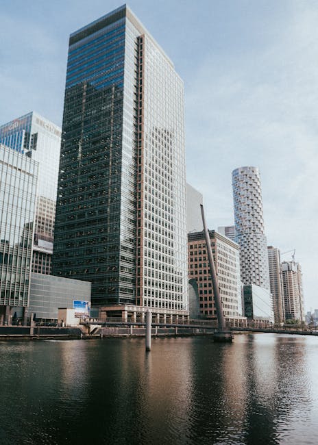 A panoramic view of Canary Wharf in London, featuring a canal running through the city with tall modern skyscrapers on either side. The buildings include glass-fronted office towers and residential bl
