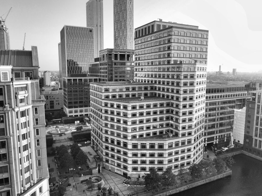 Black and white aerial photograph of a cityscape featuring modern office buildings and high-rise towers in Canary Wharf, with the foreground showing a large, multi-story building with many windows and