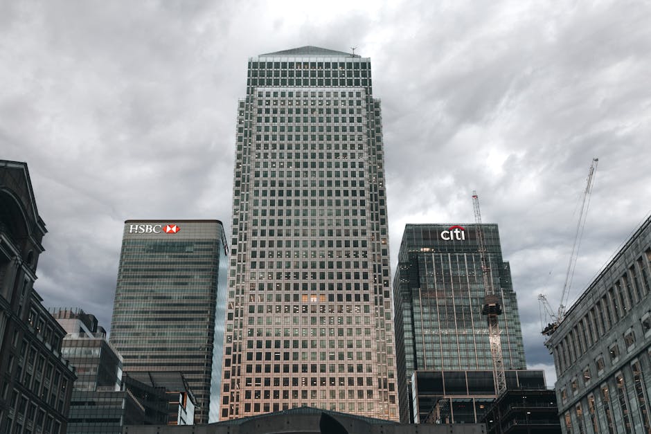 Black and white aerial photograph of a cityscape featuring modern office buildings and high-rise towers in Canary Wharf, with the foreground showing a large, multi-story building with many windows and