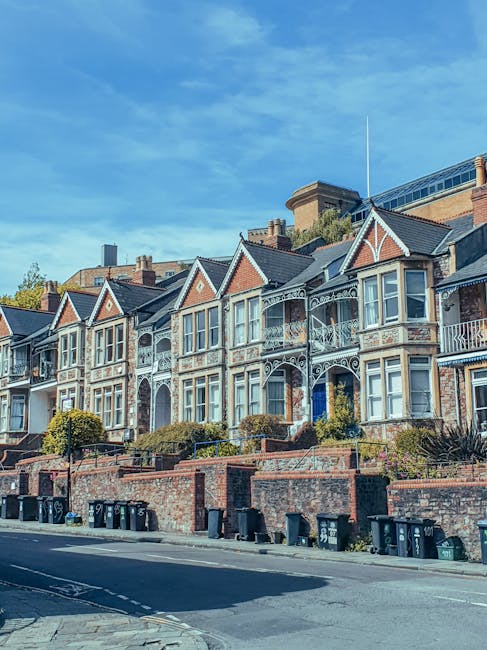 A woman with dark hair styled in a short bob is standing in the middle of a residential street with multi-storey brick buildings, some with balconies, and trees lining the pavement. She is wearing a p